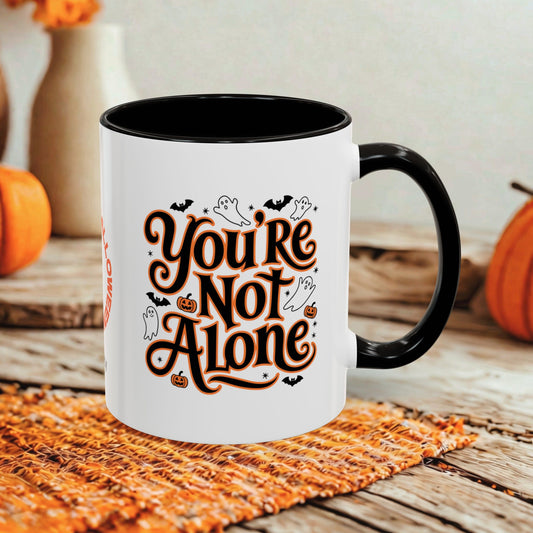 A white ceramic mug with a black handle and interior, printed with the phrase 'You're Not Alone' in stylized brown lettering with small Halloween motifs like ghosts, bats, and pumpkins. The mug sits on a woven orange placemat on a rustic wooden table, with pumpkins and a blurred background.