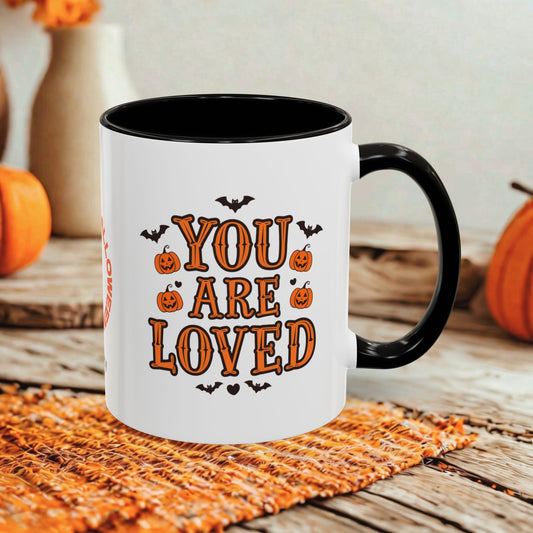A white ceramic mug with a black handle and interior, printed with the phrase 'YOU ARE LOVED' in orange letters surrounded by pumpkins and bats. The mug is styled on a rustic wooden surface with autumn decorations in the background.