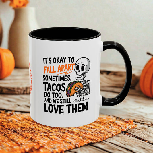 A white ceramic mug with a black handle and interior, printed with a Halloween-themed quote about fall apart tacos and a cartoon skeleton holding a taco. The mug is styled on a woven placemat with pumpkins and a neutral background.