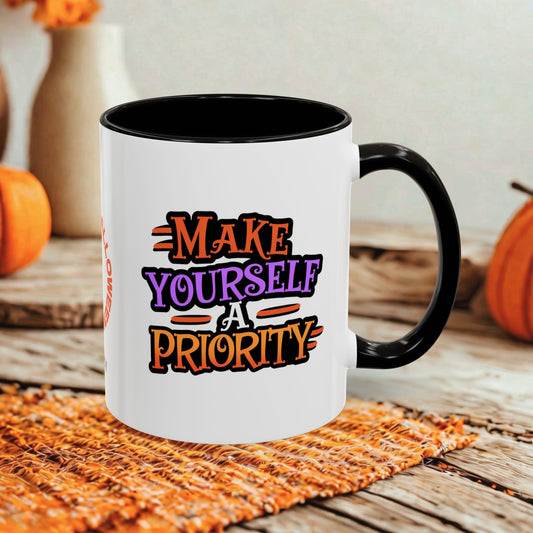 A white ceramic coffee mug with a black handle and interior, featuring the text 'MAKE YOURSELF A PRIORITY' in orange, purple, and black lettering. The mug is set on a woven orange placemat on a rustic wooden table, with pumpkins and a neutral background.