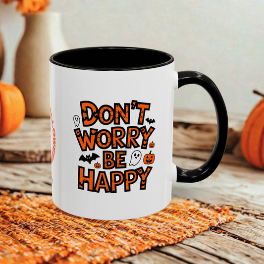 A white ceramic mug with a black interior, handle, and rim, featuring the phrase 'DON'T WORRY BE HAPPY' in orange letters with Halloween-themed illustrations. The mug is displayed on a rustic wooden table with a woven placemat and blurred orange pumpkins in the background.