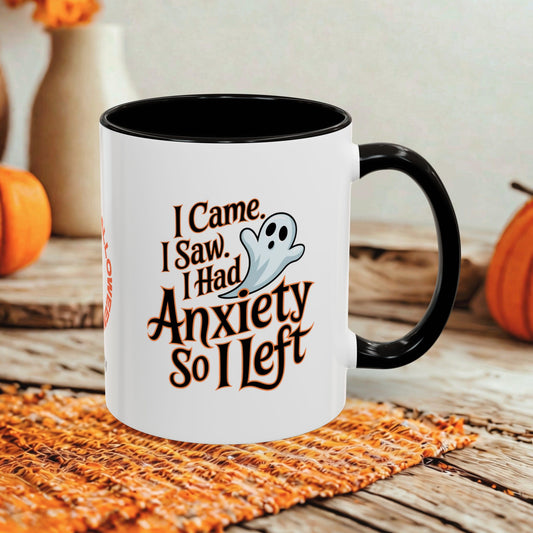 A white ceramic coffee mug with a black handle and interior, featuring a Halloween-themed graphic that reads 'I Came, I Saw, I Had Anxiety So I Left' alongside a small ghost illustration. The mug is styled on a woven orange placemat over a rustic wooden table, with pumpkins and a blurred background.