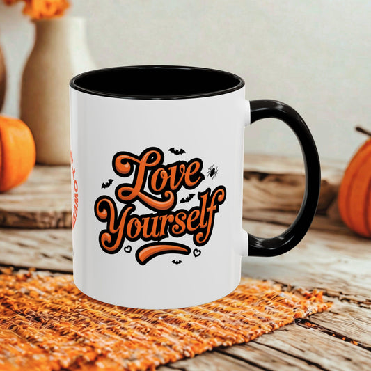 A white ceramic coffee mug with a black handle and interior, featuring the phrase 'Love Yourself' in orange script with small black bat and spider graphics. The mug is styled on a woven placemat over a rustic wooden table with pumpkins in the background.