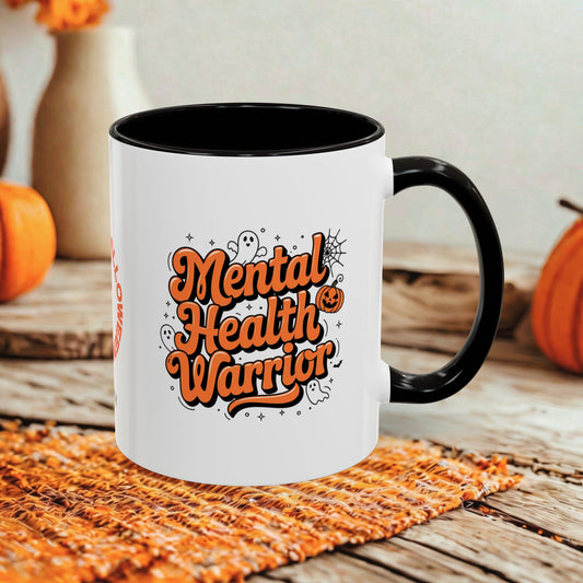A white ceramic mug with a black handle and interior, featuring the phrase 'Mental Health Warrior' in orange lettering with small decorative graphics. The mug is styled on a woven orange placemat over a rustic wooden surface, with pumpkins and a neutral wall in the background.