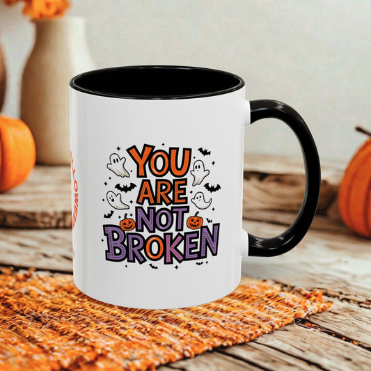 A white ceramic mug with a black handle and interior, printed with the phrase 'YOU ARE NOT BROKEN' in orange and purple lettering with cartoon ghosts, bats, and pumpkins. The mug is styled on a rustic wooden table with a woven orange placemat and blurred pumpkins in the background.