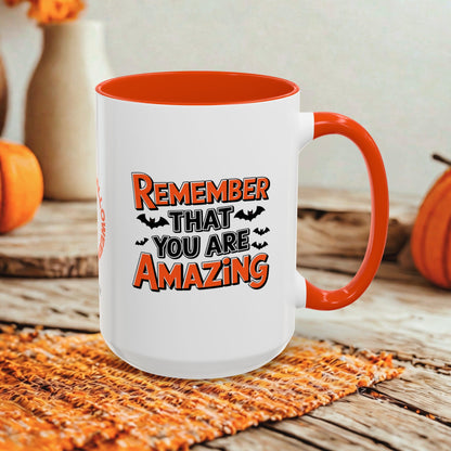 A white ceramic mug with a bright orange handle and interior, featuring the printed phrase 'REMEMBER THAT YOU ARE AMAZING' in orange and black with small bat silhouettes. The mug is styled on a woven orange placemat over a rustic wooden surface, with pumpkins and a neutral wall in the background.