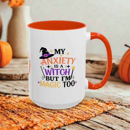 A white ceramic mug with a bright orange handle and interior, featuring a Halloween-themed printed quote that reads 'MY ANXIETY IS A WITCH BUT I'M MAGIC TOO' in orange, purple, and black lettering. The mug is styled on a rustic wooden surface with a woven orange placemat and blurred pumpkins in the background.