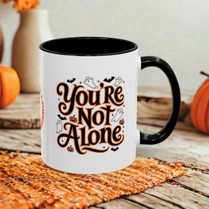 A white ceramic mug with a black handle and interior, printed with the phrase 'You're Not Alone' in stylized brown lettering with small Halloween motifs like ghosts, bats, and pumpkins. The mug sits on a woven orange placemat on a rustic wooden table, with pumpkins and a blurred background.