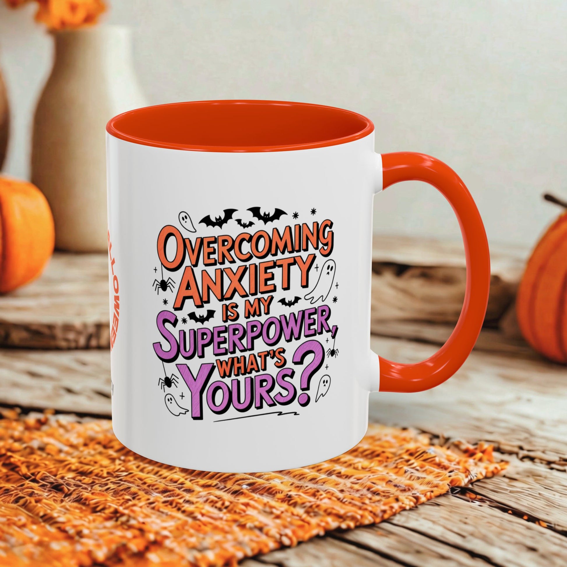 A white ceramic coffee mug with a bright orange handle and interior, featuring a Halloween-themed graphic that reads 'Overcoming Anxiety is my Superpower, What's Yours?'. The mug is styled on a rustic wooden table with a woven orange placemat, with pumpkins and a blurred background.