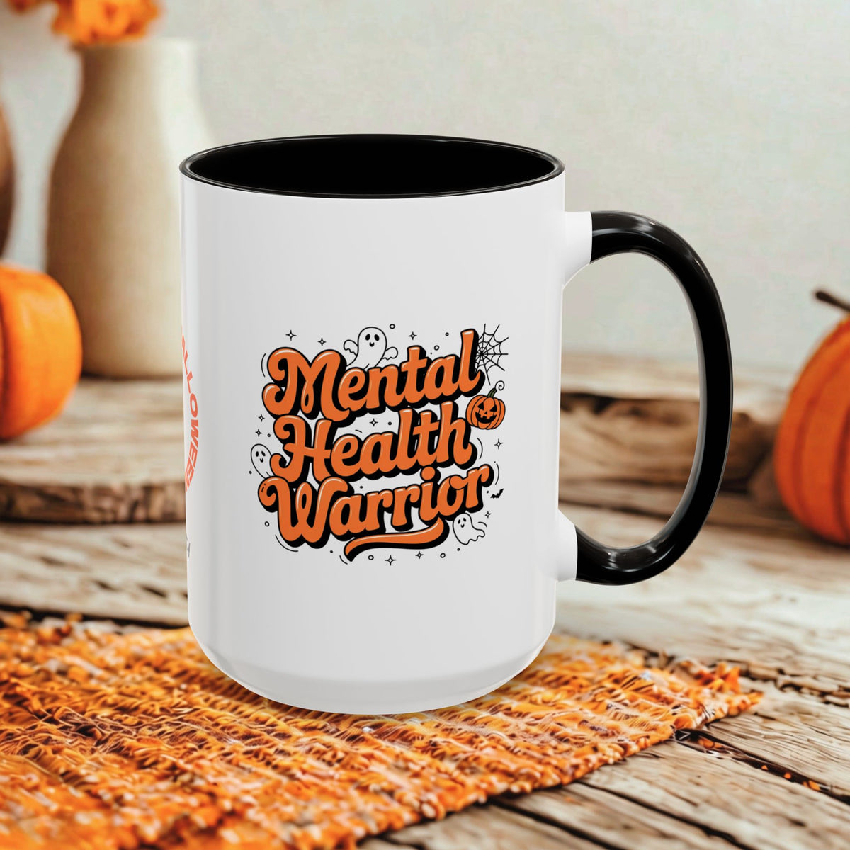 A white ceramic mug with a black handle and interior, featuring the phrase 'Mental Health Warrior' in orange lettering with small Halloween-themed doodles. The mug is styled on a woven orange placemat over a rustic wooden surface, with pumpkins and blurred decorative vases in the background.