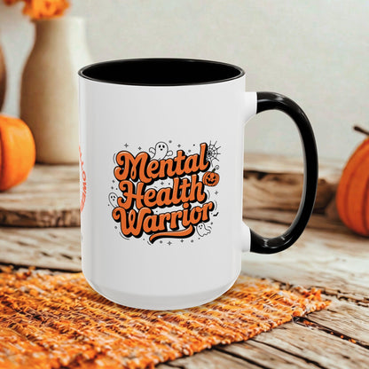 A white ceramic mug with a black handle and interior, featuring the phrase 'Mental Health Warrior' in orange lettering with small Halloween-themed doodles. The mug is styled on a woven orange placemat over a rustic wooden surface, with pumpkins and blurred decorative vases in the background.