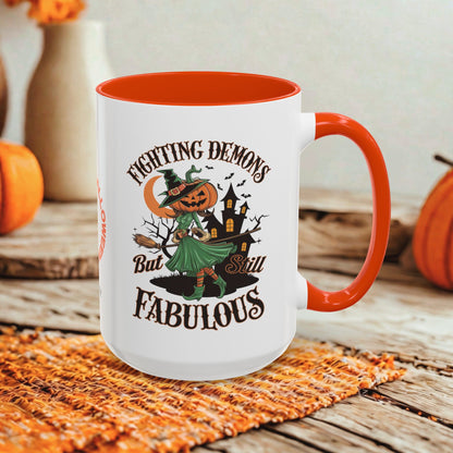 A white ceramic mug with a bright orange interior, handle, and rim, featuring a Halloween graphic of a witch fighting a pumpkin-headed demon with the text 'FIGHTING DEMONS But Still FABULOUS'. The mug is styled on an orange woven placemat on a rustic wooden table with blurred pumpkins and a vase in the background.