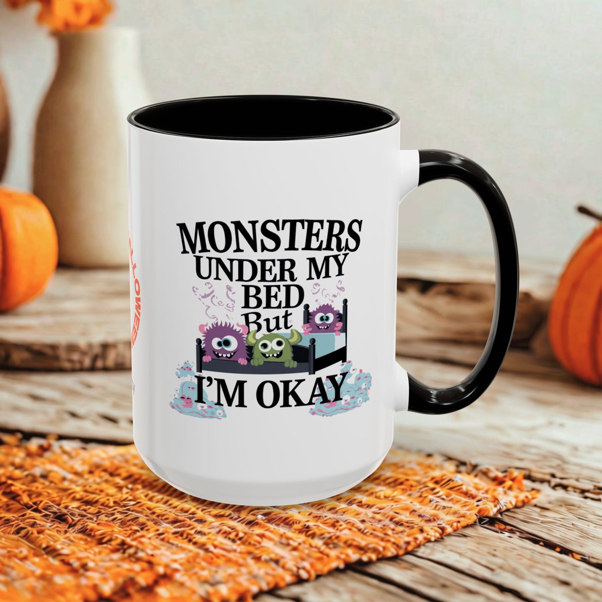 A white ceramic mug with a black handle and interior, printed with the text 'MONSTERS UNDER MY BED But I'M OKAY' and small cartoon monsters, shown on a rustic wooden surface with orange pumpkins and a woven mat in the background.