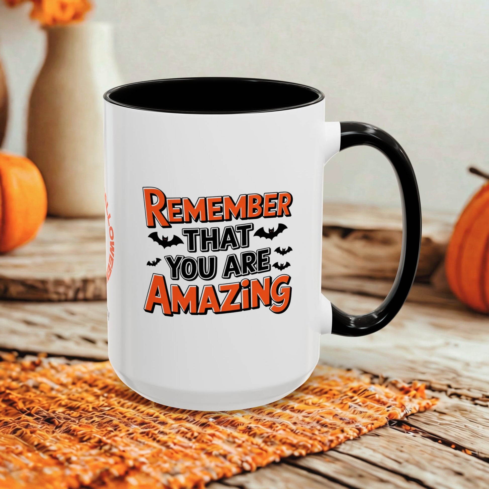 A white ceramic mug with a black handle and interior, printed with the phrase 'REMEMBER THAT YOU ARE AMAZING' in orange and black lettering with small bat silhouettes. The mug is styled on a woven orange placemat over a rustic wooden surface, with pumpkins and a neutral wall in the background.