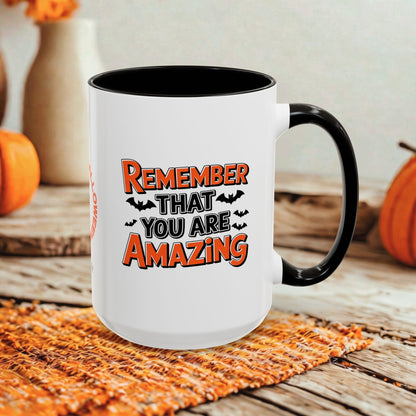 A white ceramic mug with a black handle and interior, printed with the phrase 'REMEMBER THAT YOU ARE AMAZING' in orange and black lettering with small bat silhouettes. The mug is styled on a woven orange placemat over a rustic wooden surface, with pumpkins and a neutral wall in the background.
