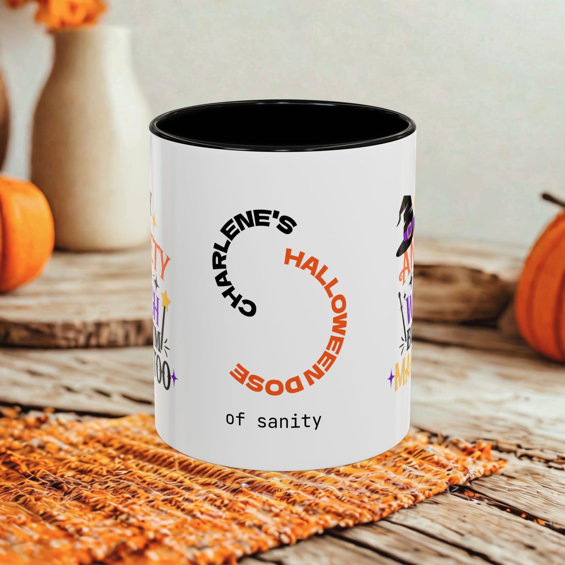 A white ceramic mug with a black interior and rim, printed with Halloween-themed text and graphics including 'CHARLENE'S HALLOWEEN DOSE of sanity' and 'of sanity' at the bottom, styled on a rustic wooden surface with orange pumpkins and a woven mat in the background.