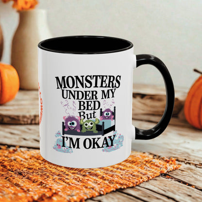 A white ceramic mug with a black interior, handle, and rim, featuring a Halloween-themed graphic that reads 'MONSTERS UNDER MY BED BUT I'M OKAY' with three colorful cartoon monsters in a bed. The mug is styled on an orange woven placemat over a rustic wooden table, with pumpkins and a blurred background.