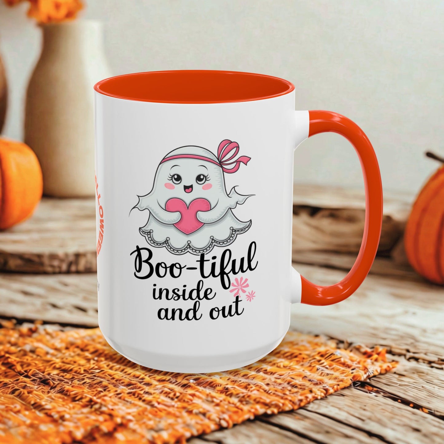 A white ceramic mug with a bright orange handle and interior, featuring a cute cartoon ghost graphic and the text 'Boo-tiful inside and out.' The mug is styled on a woven orange placemat over a rustic wooden surface, with pumpkins and blurred decorative vases in the background.