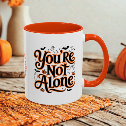 A white ceramic mug with a bright orange interior, handle, and rim, featuring the phrase 'You're Not Alone' in stylized orange and black lettering with small Halloween illustrations. The mug is set on a woven orange placemat on a rustic wooden table, with blurred pumpkins and a vase in the background.