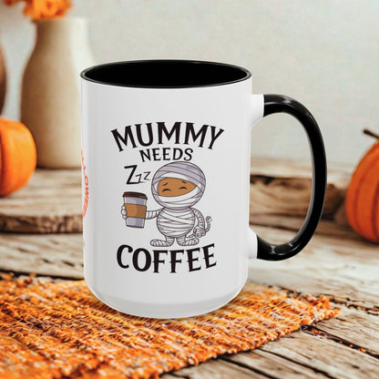 A white ceramic coffee mug with a black handle and black interior, printed with the text 'MUMMY NEEDS ZZZ COFFEE' and a cartoon mummy holding a coffee cup. The mug is styled on a woven orange placemat over a rustic wooden surface with pumpkins and a blurred background.