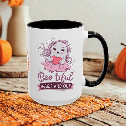 A white ceramic coffee mug with a black handle and interior, featuring a cute cartoon ghost graphic with a pink bow and the text 'Boo-tiful INSIDE AND OUT'. The mug is styled on a rustic wooden table with autumn props including pumpkins and a woven placemat.