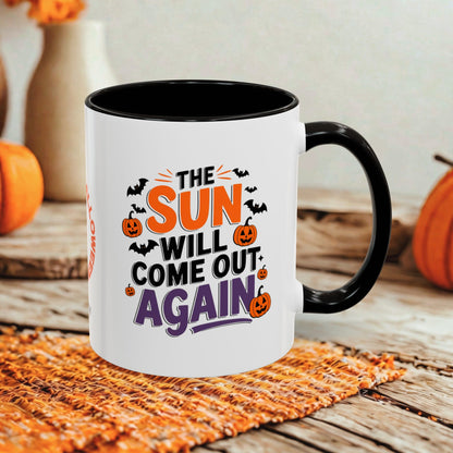 A white ceramic mug with a black handle and interior, printed with the phrase 'THE SUN WILL COME OUT AGAIN' in orange, black, and purple lettering, surrounded by Halloween graphics like pumpkins, bats, and lightning bolts. The mug is styled on a woven orange placemat over a rustic wooden table with blurred pumpkins in the background.