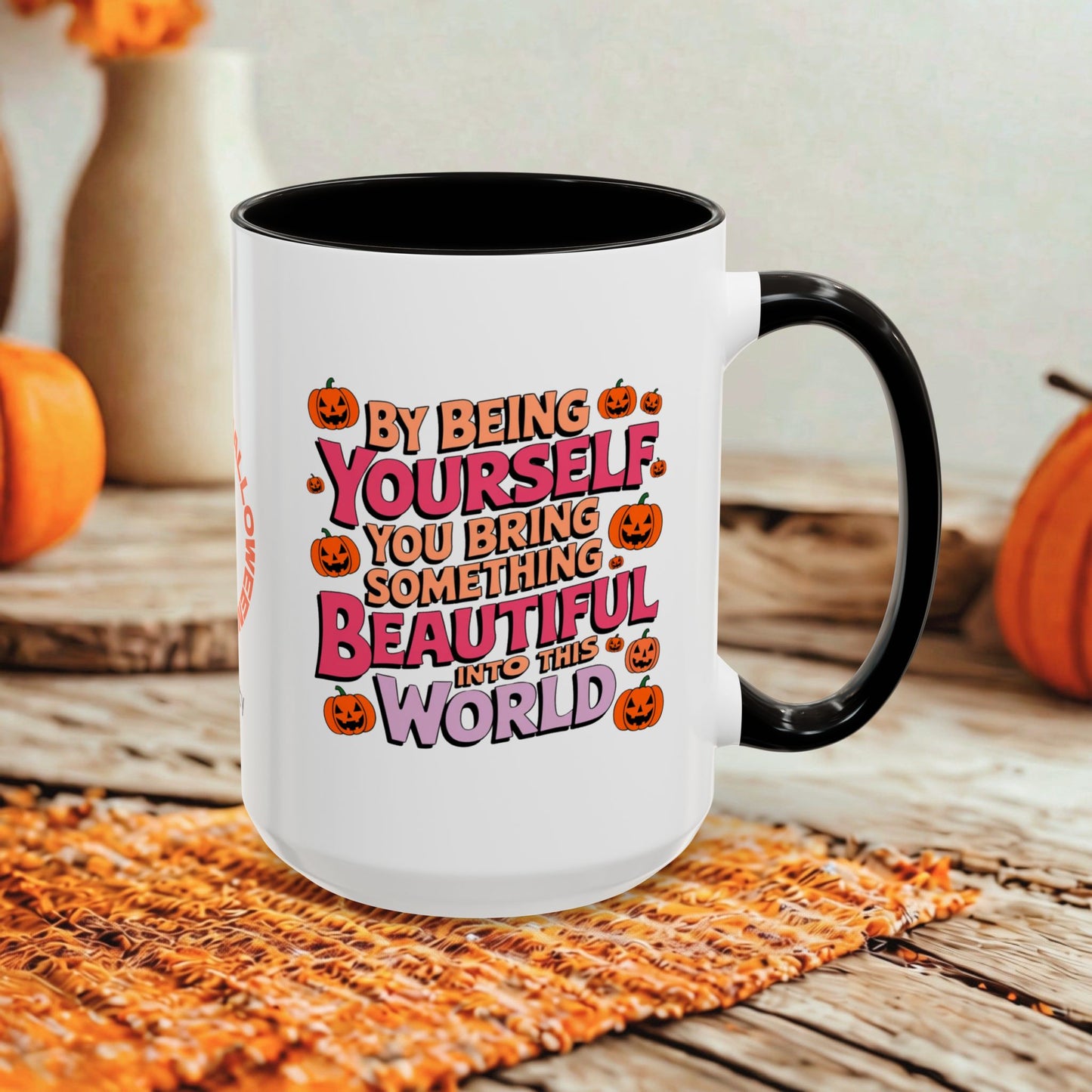 A white ceramic coffee mug with a black handle and interior, featuring a colorful Halloween-themed quote that reads 'BY BEING YOURSELF YOU BRING SOMETHING BEAUTIFUL INTO THE WORLD' surrounded by jack-o'-lanterns. The mug is set on a woven orange placemat on a rustic wooden table with blurred pumpkins and vases in the background.