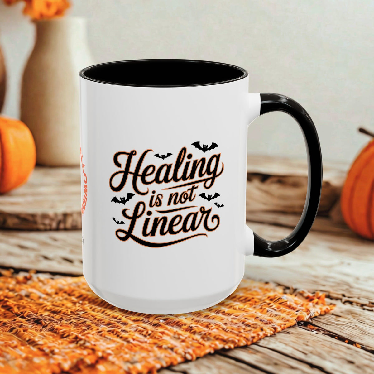 A white ceramic mug with a black handle and black interior, printed with the phrase 'Healing is not Linear' in brown script with small bat silhouettes. The mug sits on a woven orange placemat on a rustic wooden table, with pumpkins and blurred decorative vases in the background.