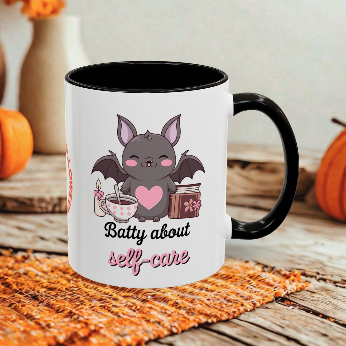 A white ceramic mug with a black handle and interior, printed with a cute bat graphic and the words 'Batty about self-care' in black and pink script. The mug is styled on a woven orange placemat over a rustic wooden table, with pumpkins and a blurred background.