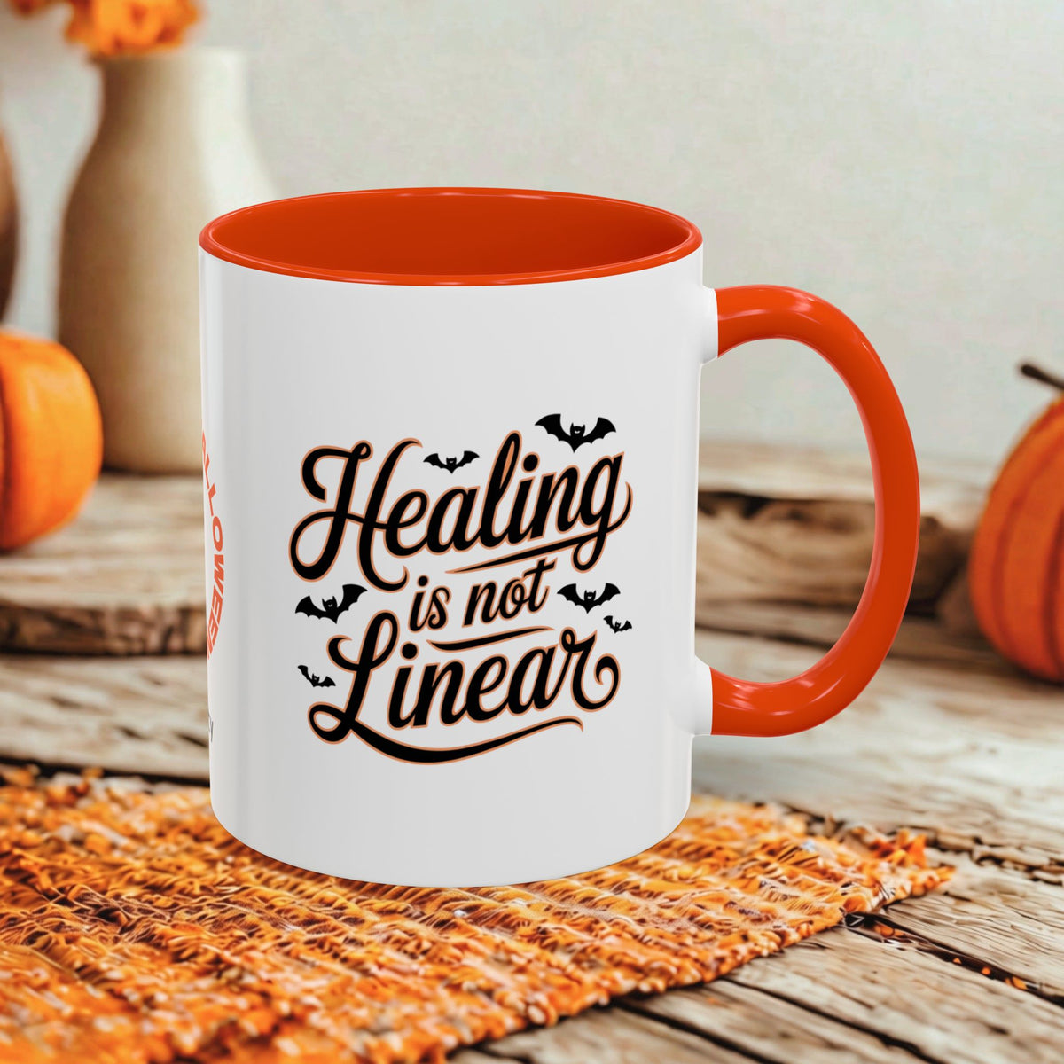 A white ceramic mug with a bright orange handle and interior, printed with the phrase 'Healing is not Linear' in dark brown script with small bat silhouettes. The mug sits on a woven orange placemat on a rustic wooden table, with blurred orange pumpkins and a neutral wall in the background.