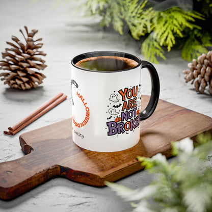 A white ceramic mug with a black handle and rim sits on a wooden cutting board, filled with a hot beverage. The mug features a Halloween-themed graphic with the text 'YOU ARE NOT BROKEN' in orange, purple, and black, alongside small cartoon ghosts and bats.