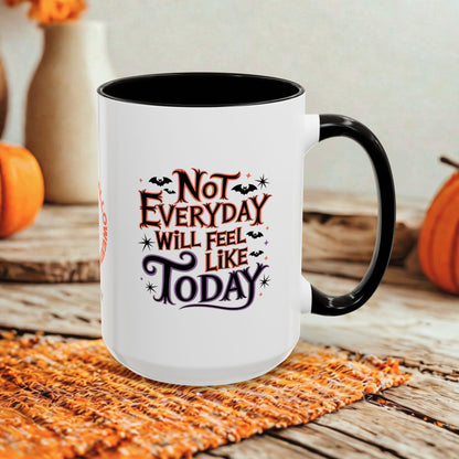 A white ceramic mug with a black handle and interior, featuring the printed phrase 'Not Everyday Will Feel Like Today' in stylized brown and purple lettering with small bat and spider motifs. The mug is photographed on a rustic wooden surface with orange pumpkins and a woven mat in the background.