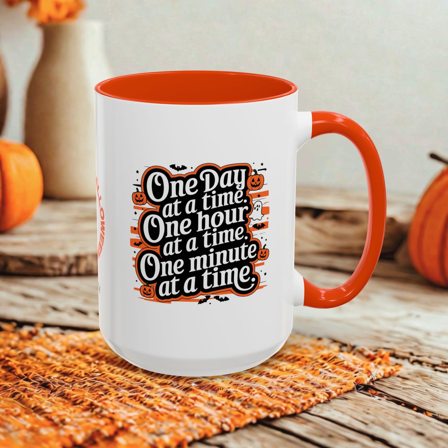 A white ceramic mug with a bright orange interior, handle, and rim, featuring a Halloween-themed printed quote that reads 'One Day at a time, One hour at a time, One minute at a time.' The mug is styled on a woven orange placemat on a rustic wooden table, with pumpkins and blurred decorative vases in the background.