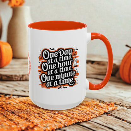 A white ceramic mug with a bright orange interior, handle, and rim, featuring a Halloween-themed printed quote that reads 'One Day at a time, One hour at a time, One minute at a time.' The mug is styled on a woven orange placemat on a rustic wooden table, with pumpkins and blurred decorative vases in the background.