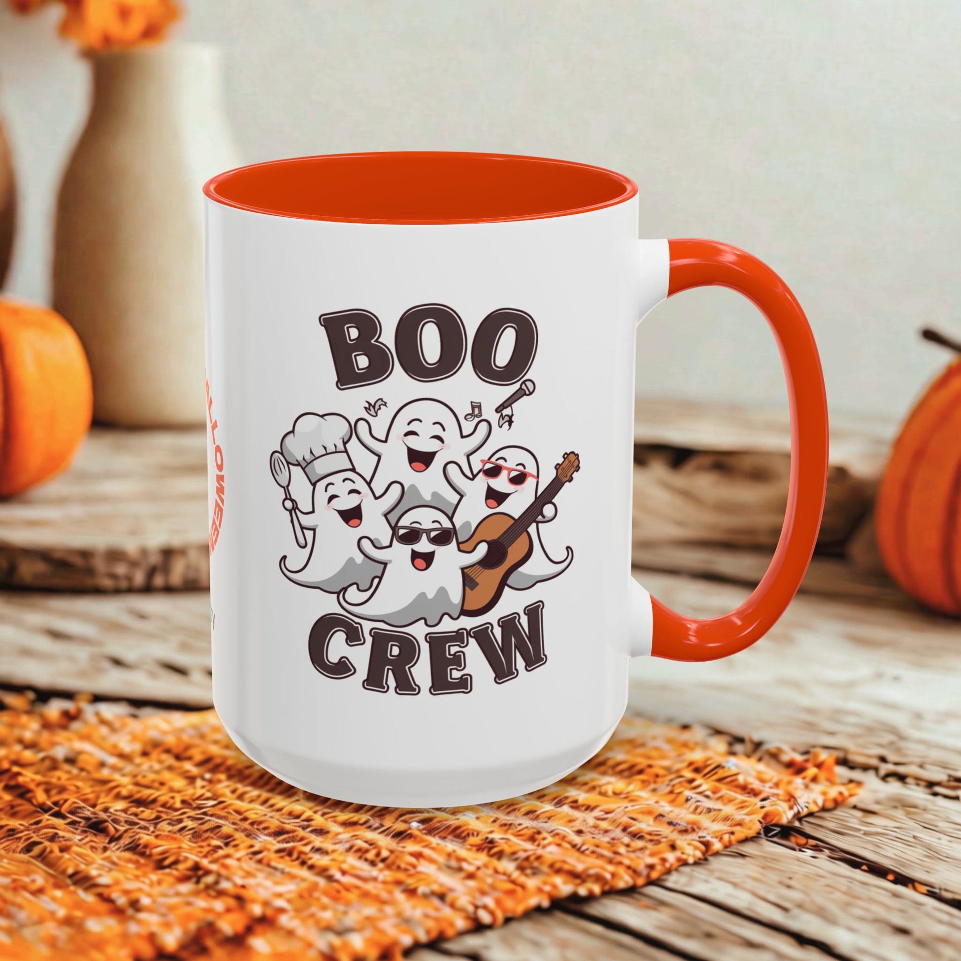 A white ceramic mug with a bright orange handle and interior, featuring a cartoon graphic of five playful ghosts and the words 'BOO CREW' in dark brown lettering. The mug is styled on a rustic wooden surface with autumn props like pumpkins and a woven placemat.