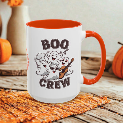 A white ceramic mug with a bright orange handle and interior, featuring a cartoon graphic of five playful ghosts and the words 'BOO CREW' in dark brown lettering. The mug is styled on a rustic wooden surface with autumn props like pumpkins and a woven placemat.