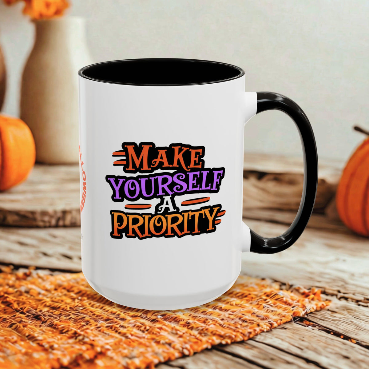 A white ceramic coffee mug with a black handle and interior, featuring the printed text 'MAKE YOURSELF A PRIORITY' in orange, purple, and black lettering. The mug is set on a woven orange placemat on a rustic wooden table, with decorative pumpkins and vases blurred in the background.