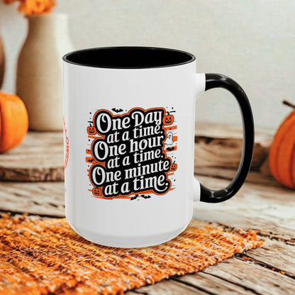 A white ceramic coffee mug with a black interior and handle, featuring a Halloween-themed printed quote that reads 'One Day at a time, One hour at a time, One minute at a time.' The mug is styled on a woven placemat with pumpkins and a neutral background.