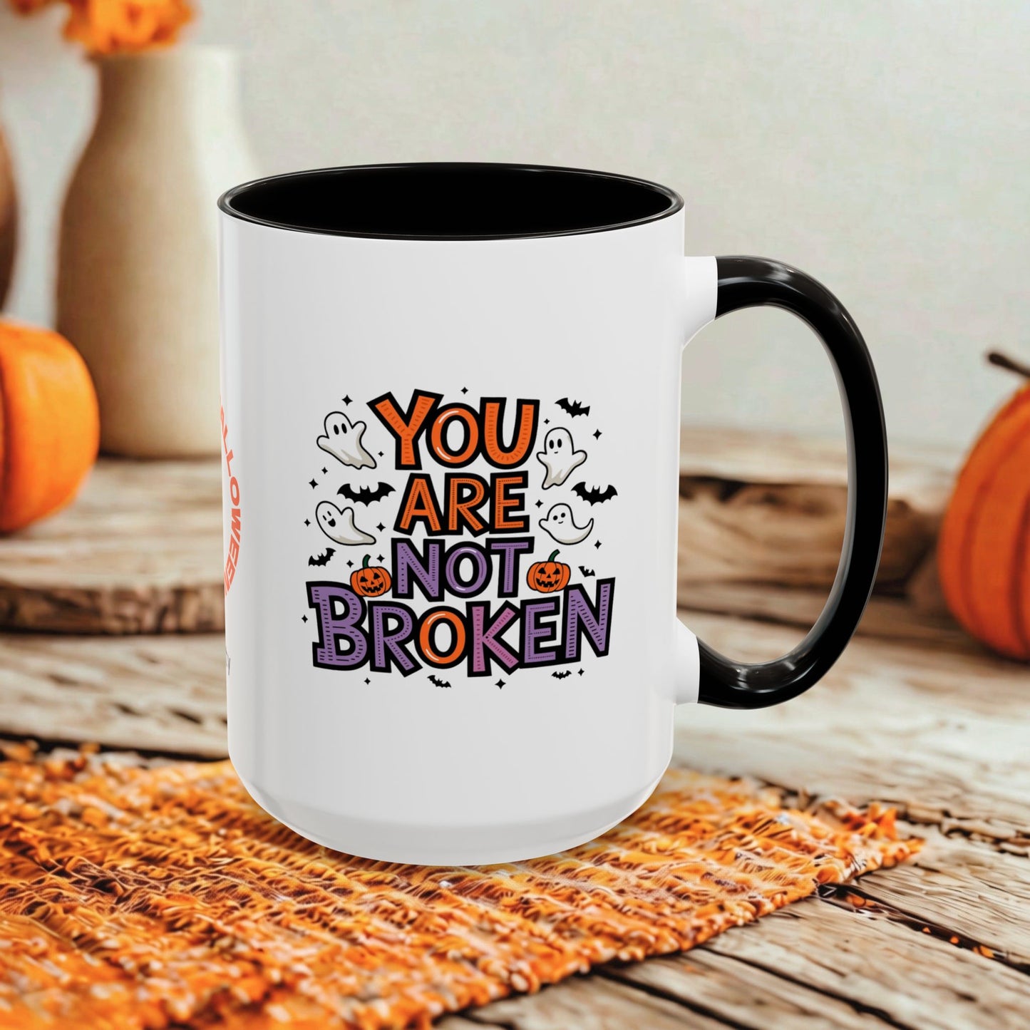 A white ceramic coffee mug with a black handle and interior, featuring a Halloween-themed graphic that reads 'YOU ARE NOT BROKEN' in orange and purple lettering with ghosts, bats, and pumpkins. The mug is set on a woven orange placemat on a rustic wooden table with blurred orange pumpkins and a beige vase in the background.