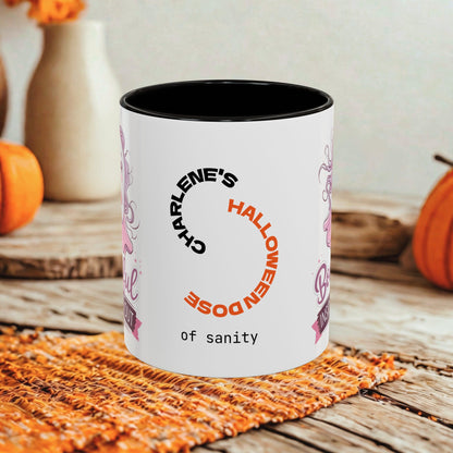 A white ceramic coffee mug with a black interior and rim, featuring a Halloween-themed graphic with the text 'CHARLENE'S HALLOWEEN DOSE of SANITY' in black and orange. The mug is set on a rustic wooden table with a woven orange placemat, with pumpkins and a vase blurred in the background.