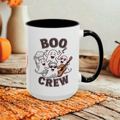 A white ceramic mug with a black handle and interior, printed with the words 'BOO CREW' and a group of smiling cartoon ghosts, shown on a rustic wooden surface with orange pumpkins and a woven placemat in the background.