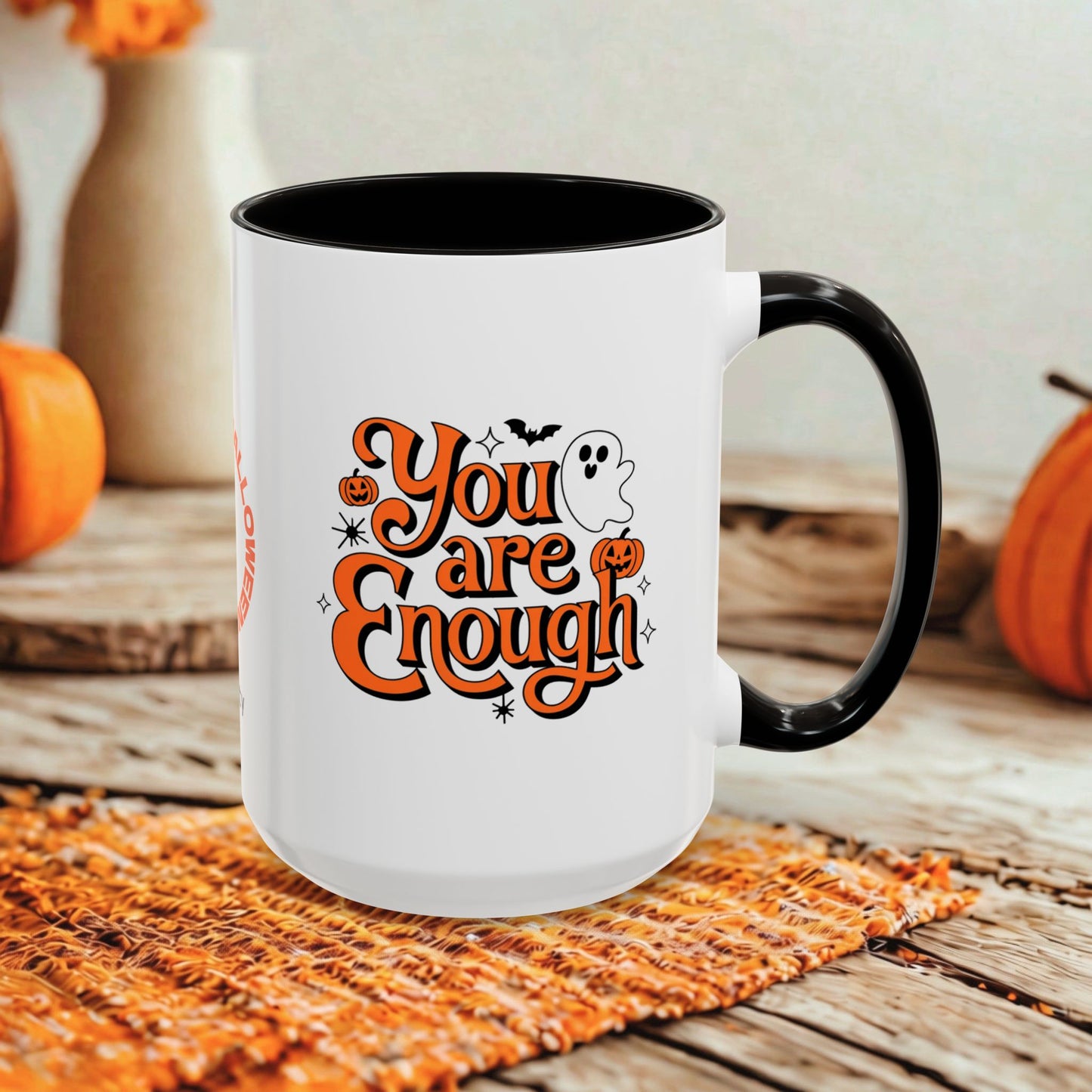 A white ceramic mug with a black handle and interior, featuring a Halloween-themed graphic that reads 'You are Enough' in orange script with pumpkins, a ghost, and bats. The mug is styled on a woven orange placemat on a rustic wooden table with pumpkins and blurred vases in the background.
