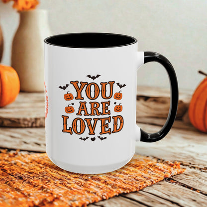 A white ceramic mug with a black handle and interior, printed with the phrase 'YOU ARE LOVED' in orange lettering with small pumpkin and bat illustrations. The mug is placed on a woven orange placemat on a rustic wooden surface, with pumpkins and a blurred indoor background.