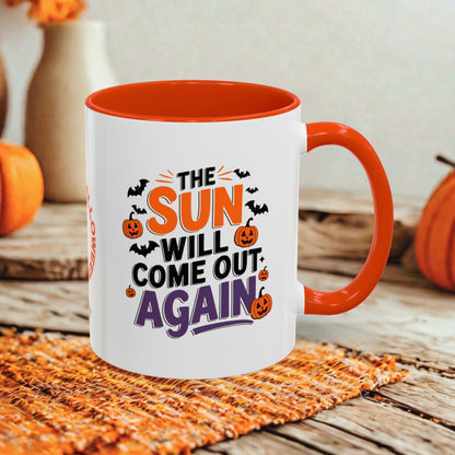 A white ceramic mug with a bright orange handle and interior, printed with the phrase 'THE SUN WILL COME OUT AGAIN' in orange, black, and purple lettering with small pumpkin and bat illustrations. The mug sits on a woven orange placemat on a rustic wooden table, with blurred pumpkins and a vase in the background.