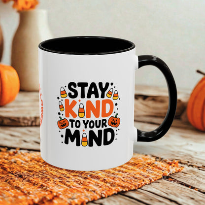 A white ceramic mug with a black handle and interior, printed with the phrase 'STAY KIND TO YOUR MIND' in black and orange lettering with Halloween-themed candy and pumpkin illustrations. The mug is photographed on a rustic wooden surface with orange woven placemat and blurred pumpkins in the background.