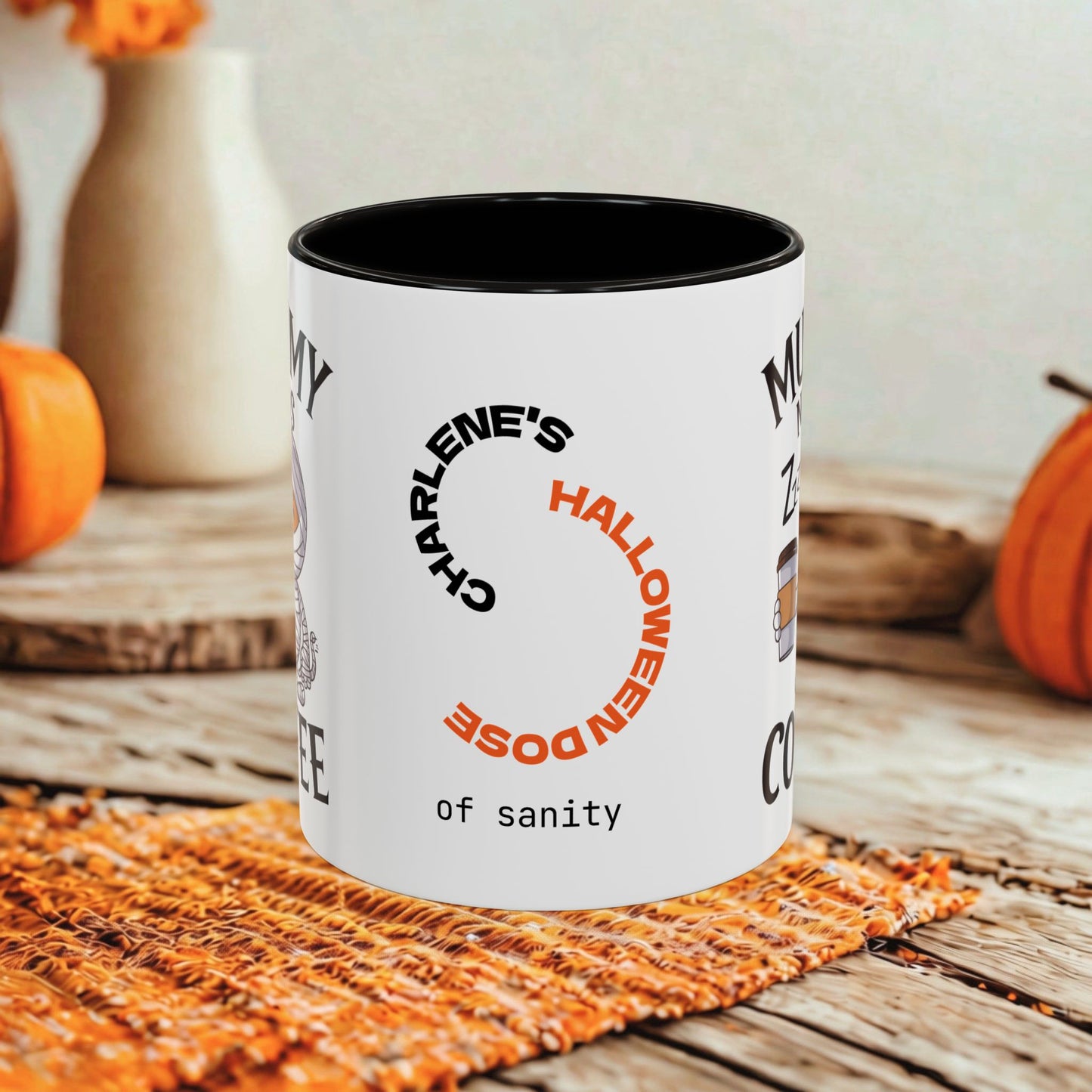 A white ceramic coffee mug with a black interior and rim, featuring a circular graphic that reads 'CHARLENE'S HALLOWEEN DOSE of SANITY' in black and orange lettering. The mug is set on a rustic wooden table with a woven orange placemat, with pumpkins and a vase blurred in the background.