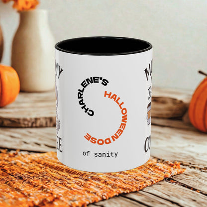 A white ceramic coffee mug with a black interior and rim, featuring a circular graphic that reads 'CHARLENE'S HALLOWEEN DOSE of SANITY' in black and orange lettering. The mug is set on a rustic wooden table with a woven orange placemat, with pumpkins and a vase blurred in the background.