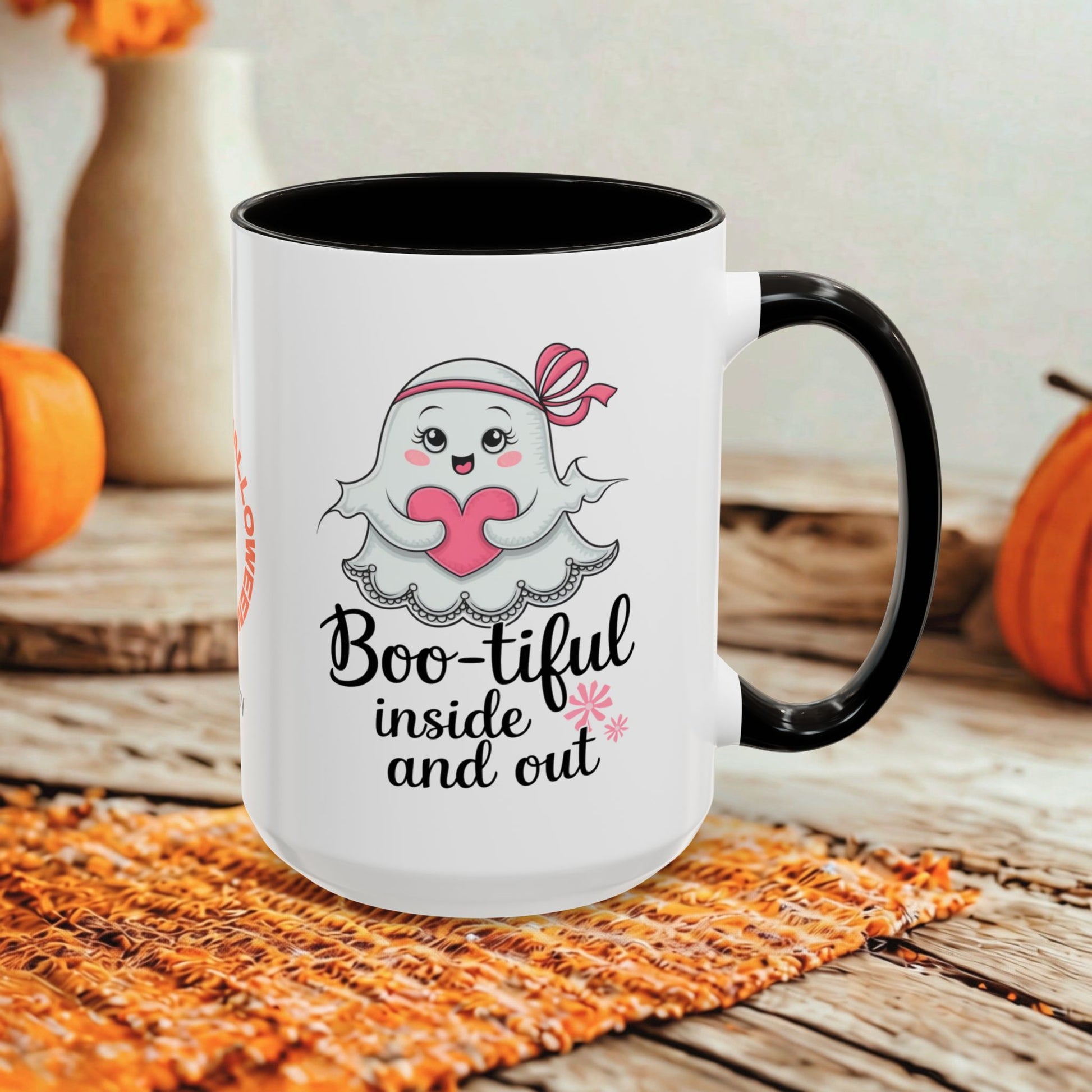A white ceramic mug with a black handle and black interior, featuring a cute cartoon ghost holding a pink heart and the text 'Boo-tiful inside and out.' The mug is styled on a woven orange placemat over a rustic wooden surface with pumpkins and blurred decorative vases in the background.