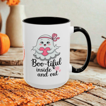 A white ceramic mug with a black handle and black interior, featuring a cute cartoon ghost holding a pink heart and the text 'Boo-tiful inside and out.' The mug is styled on a woven orange placemat over a rustic wooden surface with pumpkins and blurred decorative vases in the background.