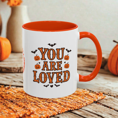 A white ceramic mug with a bright orange handle and interior, printed with the phrase 'YOU ARE LOVED' in orange letters surrounded by small black bats and pumpkins. The mug is styled on an orange woven placemat over a rustic wooden surface with seasonal pumpkins and a vase in the background.